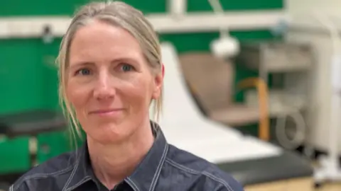 Dr Nicki Mazey sits in a doctor's room in front of a examination bed. She has blonde hair that is tied back and wears a dark shirt with collar.