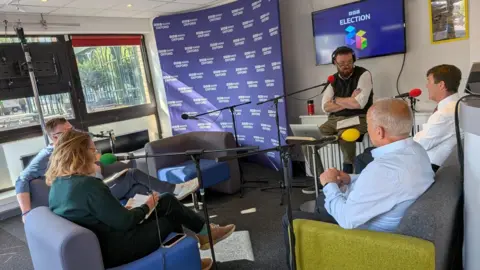 Four people sat on sofas in front of microphones. The presenter is sat on a chair. Behind him is purple BBC election branding.