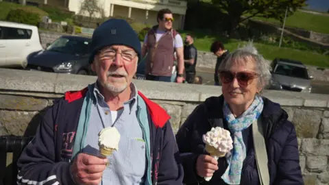 A couple sitting eating large ice creams. Both are wearing jackets. Dave has a beanie hat while June is wearing sunglasses