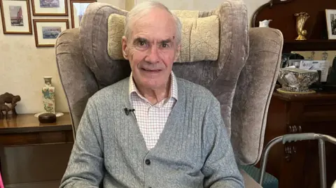 John Barrington Powell, 86, sitting in a chair at home, wearing a grey cardigan and light coloured shirt, and smiling at the camera, with a walking frame nearby 