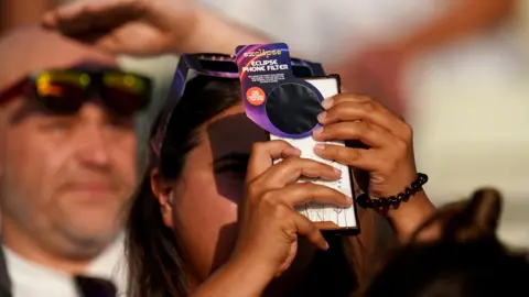 PA Person holding a mobile phone fitted with an eclipse phone filter in the foreground, aiming it upwards. Another person nearby wears eclipse-viewing glasses and shades their eyes with a raised hand. The scene is outdoors in bright sunlight, with a crowd visible in the background.