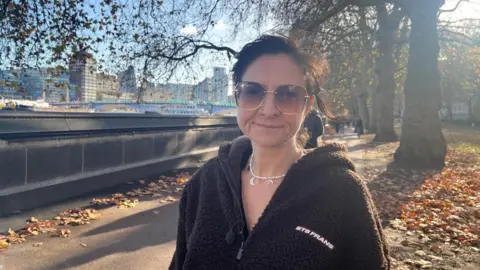 Jess Miller is standing next to the embankment in London. You can see the River Thames over her right shoulder and a row of trees directly behind her. She has short dark hair and wearing large brown sunglasses. She is wearing a silver necklace and a dark brown hoody