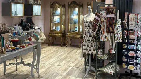 Seb Cheer/BBC The interior of a shop showing wooden flooring and a table of jewellery and gifts, and some gold display cabinets filled with items in the background