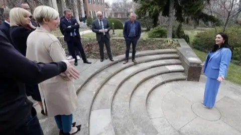 Emma Little-Pengelly standing in a circle with round steps leading down to it. She has long dark hair and wearing a light blue suit. Several members of the press are standing on the steps looking down towards her.