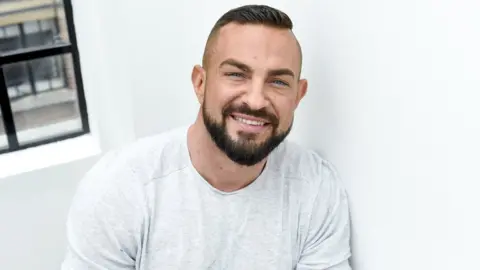 James Watkins/BBC Robin Windsor poses against a white wall, there is a corner of a window in the top left of the photo. He is smiling and wearing a white long sleeved t-shirt.
