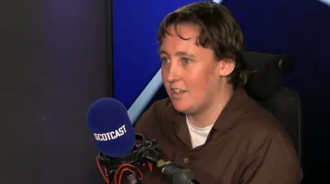 Mhairi Black sitting in radio studio behind blue microphone with Scotcast on it. She has short brown hair, and is wearing a brown shirt over a white T-shirt.