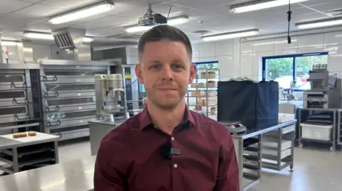 Kate Bradbrook/BBC Simon Curran, wearing a burgundy shirt, standing in a kitchen, in a mill, with metals machinery behind him. 