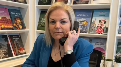 Linda Hill Miller, a woman with blonde, shoulder length hair. She is wearing a black top with a blue blazer and holds a phone to her ear. In the background there are travel brochures for different countries on shelves.