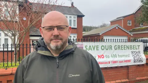Photograph of Syd Cottle from Save Winstanley Green Belt. The image shows a Save Our Greenbelt campaign poster in the background.