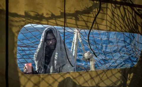 Getty Images An African man with a grey blanker over his head is looking straight into the camera and holding a cup on board a ship anchored off Sicily, Italy - Saturday 26 January 2019