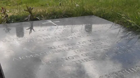A large flat headstone lays on the ground, surrounded by grass which pokes over it's edges. It's a black marble like stone with a cross at the top. Only some text is visible, showing the name 'William George Neely' and his age, of 77. Below it reads 'Rector of Keady'. 