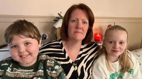 Shows a woman in a black and white zebra-patterned top with a young boy in a Christmas jumper to the left and girl with fair hair in light-coloured Christmas jumper to the right.