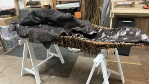 Lizzy Steel / BBC A woolly, black and white burial shroud sits in a wicker basket in the artist's workshop. It is displayed on a white wooden stand. 