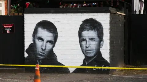 Emma Gibbs A black-and-white mural of Liam and Noel Gallagher painted on a brick wall behind a traffic cone and a yellow plastic strip