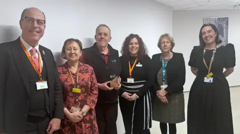 Paul Newman is wearing a black bodywarmer over a claret polo shirt. He is smiling and holding his award. There are three women to his left and a man and a woman to his right. They are all smartly-dressed and smiling.