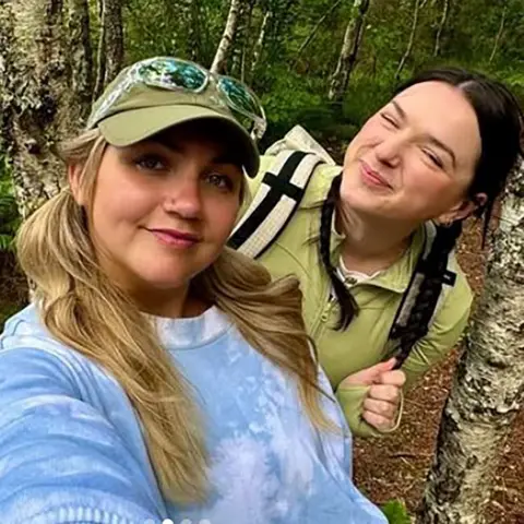 Emily & Lucy Emily and Lucy - 2 women opinionated successful a forested area, surrounded by trees. One is wearing a ray bluish tie-dye garment and has agelong blonde hair. The different is dressed successful a greenish garment and carrying a backpack. 
