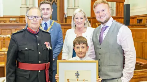Middlesbrough Council Alan Peacock's family standing in the council chamber which is made up of dark wood seating. Diane Symington has shoulder-length blonde hair and is wearing white, stud earrings and a white jacket. Luca stands in front of her holding a large certificate in a gold frame. He has brown hair and is wearing a white shirt. Harry stands to the right and has blonde hair, a darker beard and a grey waistcoat with a purple tie and pink shirt. They are all smiling into the camera. Chris Cooke stands on the left in a blue suit with a gold mayoral necklace on. A man stands slightly in front of him in black and red military clothing.