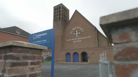 A church building with a large bell tower to the left hand side, and a steep pitched roof can be seen between two red-brick pillars. It has three arched entrances to the front, but two of them are closed off by blue shutters. Large letters on the side of the building read 'St Brendan's' and below is a quote reading "I am the way, the truth and the life" Jesus Christ. 