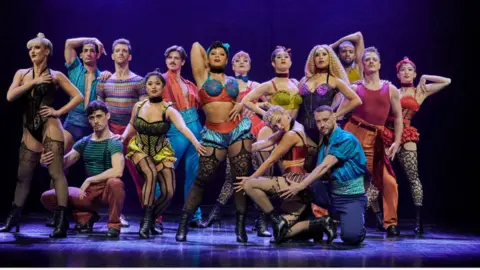 Matt Crockett The cast of Moulin Rouge posing on a dark stage, wearing scantily-clad costumes with the ladies in corsets of bright colours and lacy stockings and the men in coloured trousers and vests or shirts. The performers at the front have their hands on each others' legs in suggestive poses.