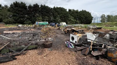 Sarah-May Buccieri / BBC View of the aftermath of the fire with the charred remains of several structures and burnt furniture and electrical equipment on the ground. In the distance is a van and caravan parked by a row of trees