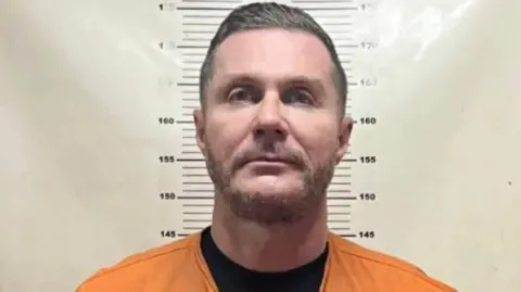 Police mugshot of Steven Lyons. He is standing against a white height chart and staring directly at the camera. He has heavy stubble and short dark hair, combed in a side shed. He is wearing a black t-shirt and orange prison overalls. 