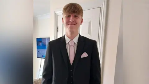 Family Handout 18-year-old Alfie Gray wears a suit with a pink tie and is smiling down the camera.