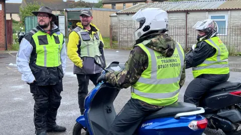 Two men in motorbike leathers and wearing high-viz yellow jackets stand facing two other men who are similarly dressed but also on scooter-style motorbikes. The men on the bikes are also wearing crash helmets
