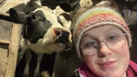 BBC Mary in a winter hat with a cow right behind her in a pen