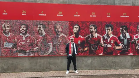 Igor Gois Igor stands on a paved area in front of a large red mural showing football players in matching kits. Above the mural, large letters spell “LISBOA” on a concrete wall, with player names and numbers displayed along the top.