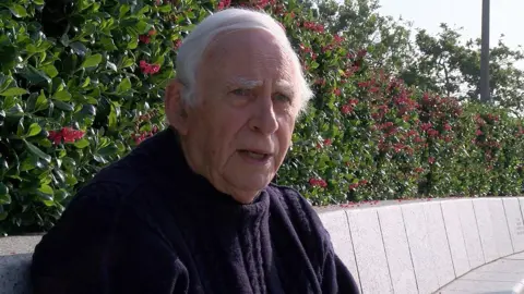 David sits on a white granite curved bench seat. He has short white hair and is wearing a navy blue knitted jumper.