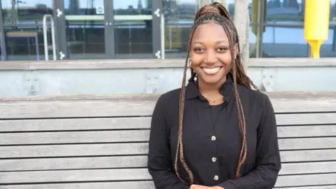 University of East London A black woman with braids, wearing a black shirt and trousers, smiles towards the camera as she sits on a wooden bench