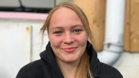 Christine Butler/BBC Lillah is wearing a black hoodie and smiling at the camera. She has blonde hair.