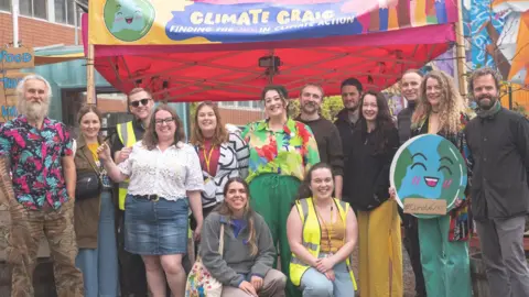 14 people stand under a yellow banner which reads 'Climate Craic.' Behind the banner are grey-bricked buildings 