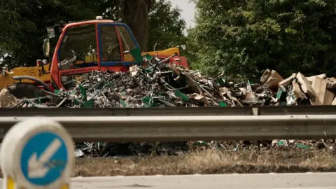 DJ McLaren/BBC A pile of scrap material next to the road, with a small digger and some trees in the background.