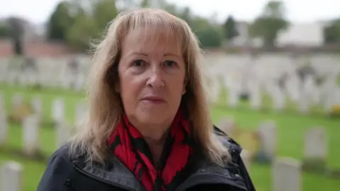 A woman with long blonde hair and a scarf decorated with red poppies is stood in front of rows of white graves in a green cemetery.