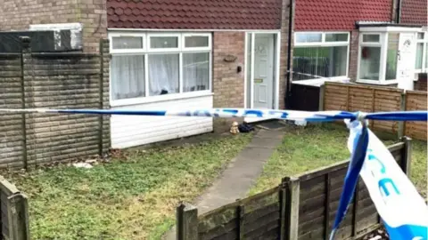 A house with police tape in front of it, with a small garden laid to lawn, with a diagonal path leading to a white front door, which is open. 