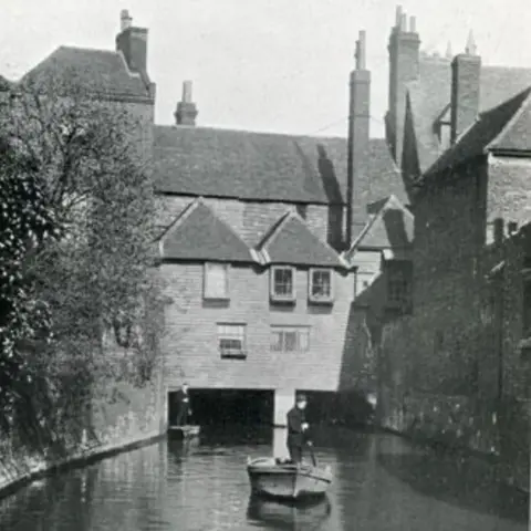 Eastbridge Hospital A medieval building bridging over a river, in black and white. Two punters are seen on the water, with trees on the wall banking the water.