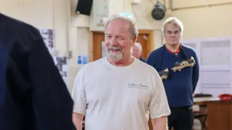 Clara Mbirini Mullan smiles at another actor we see from behind in a bright rehearsal room. Another man, with football boots around his neck, stands behind him while watching on. Mullan is wearing an off-white t-shirt with cursive script on the left side.