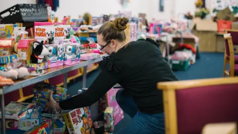 The Salvation Army A woman wearing a black top and jeans holding several boxes of toys in one hand. The table in front of her is full of toys. 