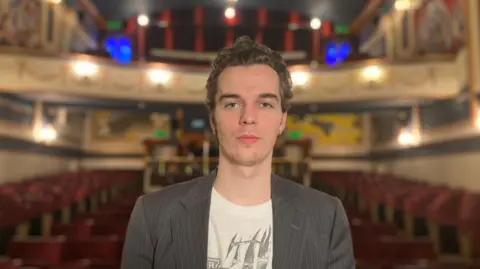 Bridport Arts Centre George Earwicker looking at the camera, has dark hair, blue eyes and is wearing a suit jacket.