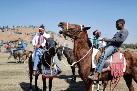 AFP Men and a boy on brown horses. In the background there are hills and a crowed of people scattered around.