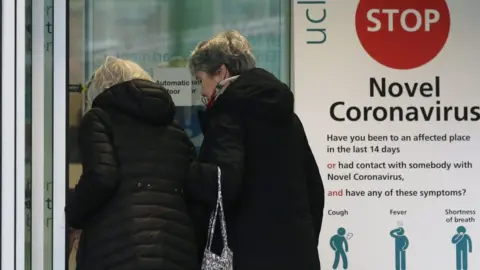 Getty Images Two women walk past a sign providing guidance information about novel coronavirus (COVID-19) at one of the entrances to University College Hospital in London on March 5, 2020. - The number of confirmed cases of novel coronavirus COVID-19 in the UK rose to 85 on March 4, with fears over the outbreak delaying the global release of the new James Bond movie and causing lack of demand for air travel that has proved the final nail in the coffin for British regional airline Flybe which went into administration on March 5.