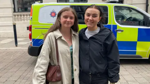 Two young women with brunette hair are posing for a picture on a street in front of a police van.
