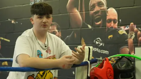 He is wearing a white pattered T'shirt and leaning on the boxing ring ropes. He has short brown hair.