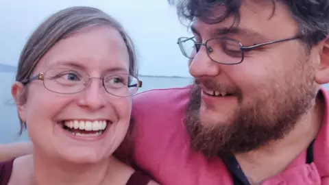 Alyssa and her husband are hugging, with a coastal view behind them. They are both smiling and she is looking off to the right, while he is looking at his wife.