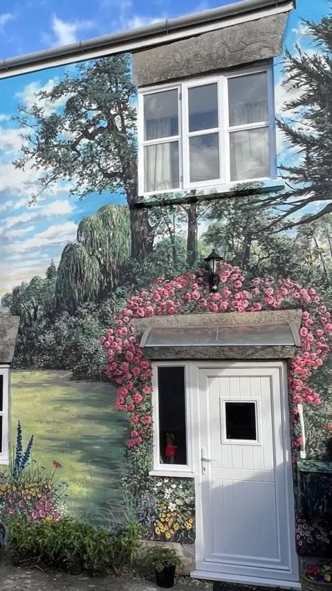 Mural on exterior wall of a home, showing a blue sky, trees and a bush with pink flowers above the white front door.