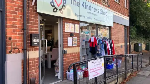 Two-storey brick building, with the ground floor operating as a shop. There is a rail of clothes outside the shop and toys on display in the window. A sign on the building says "Kindness Foundation Community Hub". There is a ramp up to the door with a black railing.