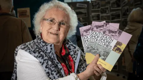 Blackpool Council Elaine Smith holds copies of the book celebrating South Shore. She has white hair, spectacles, wears a red patterned scarf and has a zip up cardigan with white sleeves and grey and black leopard skin patterned collar.