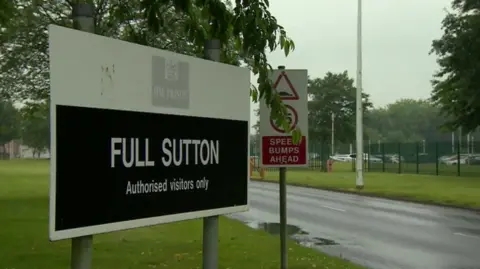 Sign at the entrance to HMP Full Sutton displaying the words “FULL SUTTON” and “Authorised visitors only”. The sign stands next to a road bordered by grass and trees. A fence and prison grounds are visible in the background under an overcast sky.