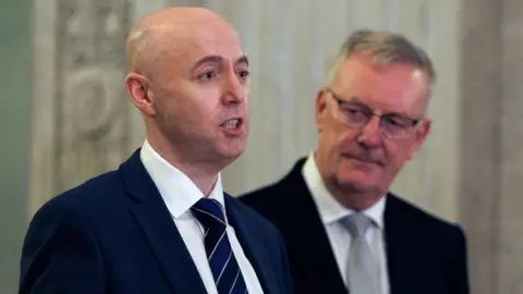 PA Media Jon Burrows standing beside Mike Nesbitt. Burrows is bald and is wearing a white shirt, navy tie and navy suit. Nesbitt has grey hair and i wearing a white shirt, grey tie and black suit. They are standing in front of the staircase in Stormont Buildings. 